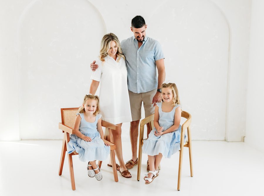 A family of four with the girls seated in wooden chairs in front of the white arch
